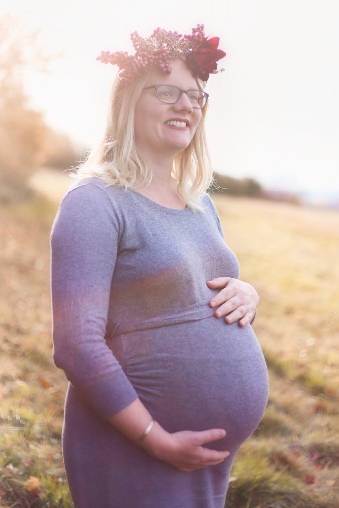 Eine schwangere Frau mit Blumenkranz steht im Grünen und hält lächelnd ihren Babybauch