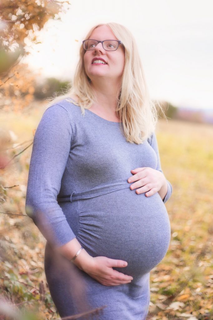 Eine schwangere Frau mit steht im Grünen und hält lächelnd ihren Babybauch
