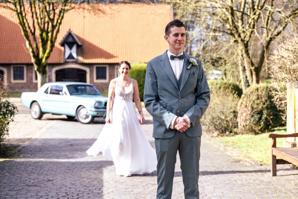 Foto eines Brautpaares am Hochzeitstag in dem die Braut sich für ein erstes Treffen von hinten dem Bräutigam nähert.